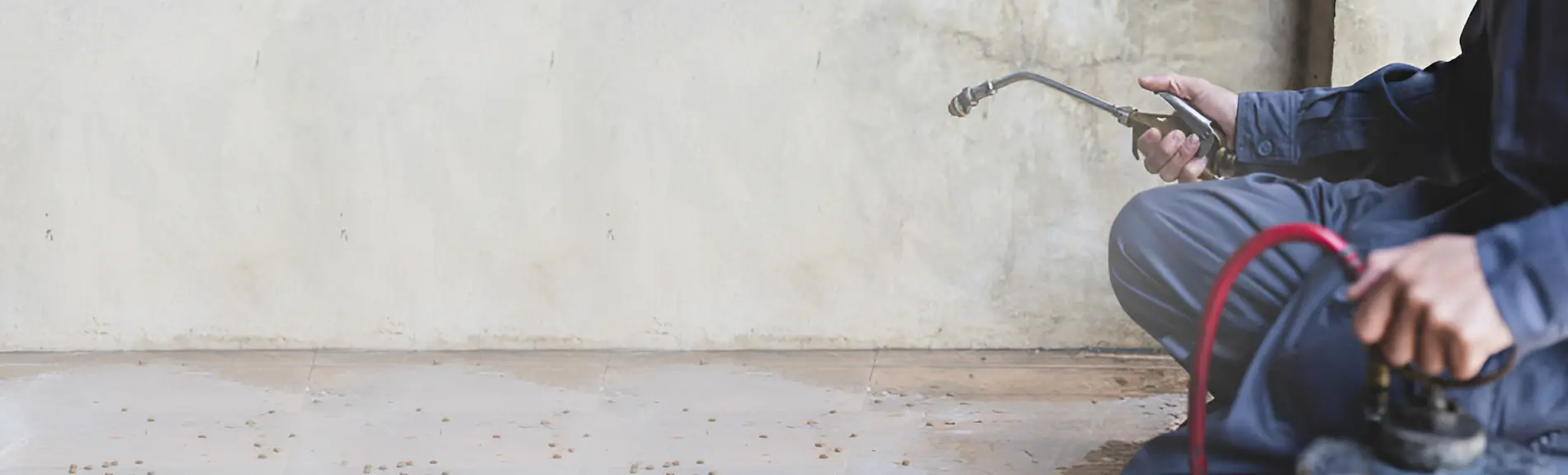 A person sitting cross legged, their body cropped out of the image. They are facing the center of the image with a sprayer in their right hand spraying a concrete wall and floor.